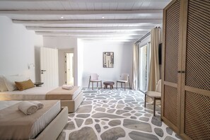 Una camera d'albergo moderna con pavimento effetto pietra, un letto con biancheria beige, un'area salotto con tavolo e sedie, e un'ampia finestra con tende.