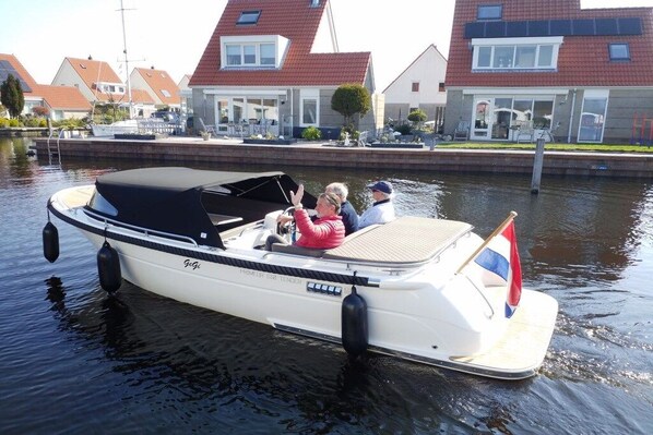 Un étroit canal, sur lequel navigue un petit bateau blanc surmonté d’un auvent noir. Deux personnes se trouvent à bord, et on aperçoit, au fond, des maisons aux toits rouges.