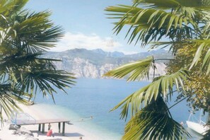 A beach with clear blue water, palm trees, and a rocky mountain backdrop. - Holiday apartment with 2 bedrooms (Malcesine)