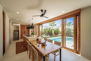 A modern kitchen with a central island, a dining area with a view of a swimming pool, and a ceiling fan. - Villa GIGI Balangan (Balangan)