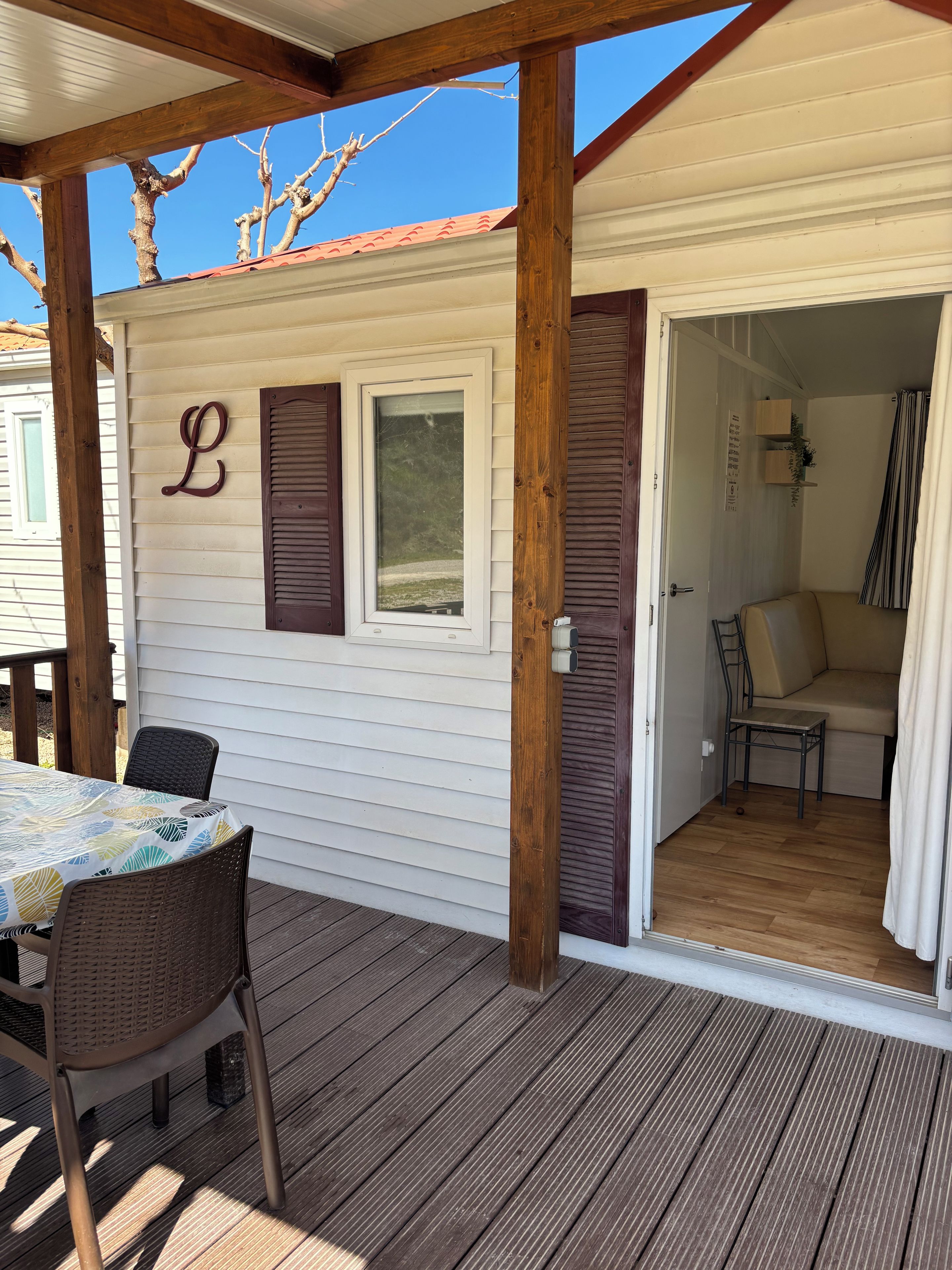 Un espacio exterior techado con una mesa y sillas, una terraza de madera y una puerta que da a un ambiente interior con un sofá y una mesita.