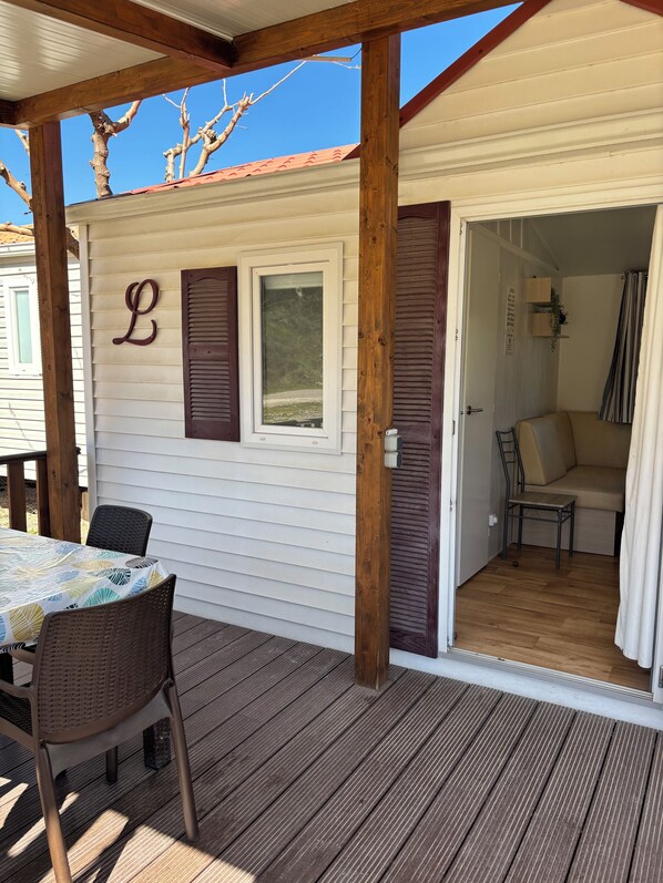 A covered outdoor area with a table and chairs, a wooden deck, and a door leading to an interior space with a sofa and a small table.