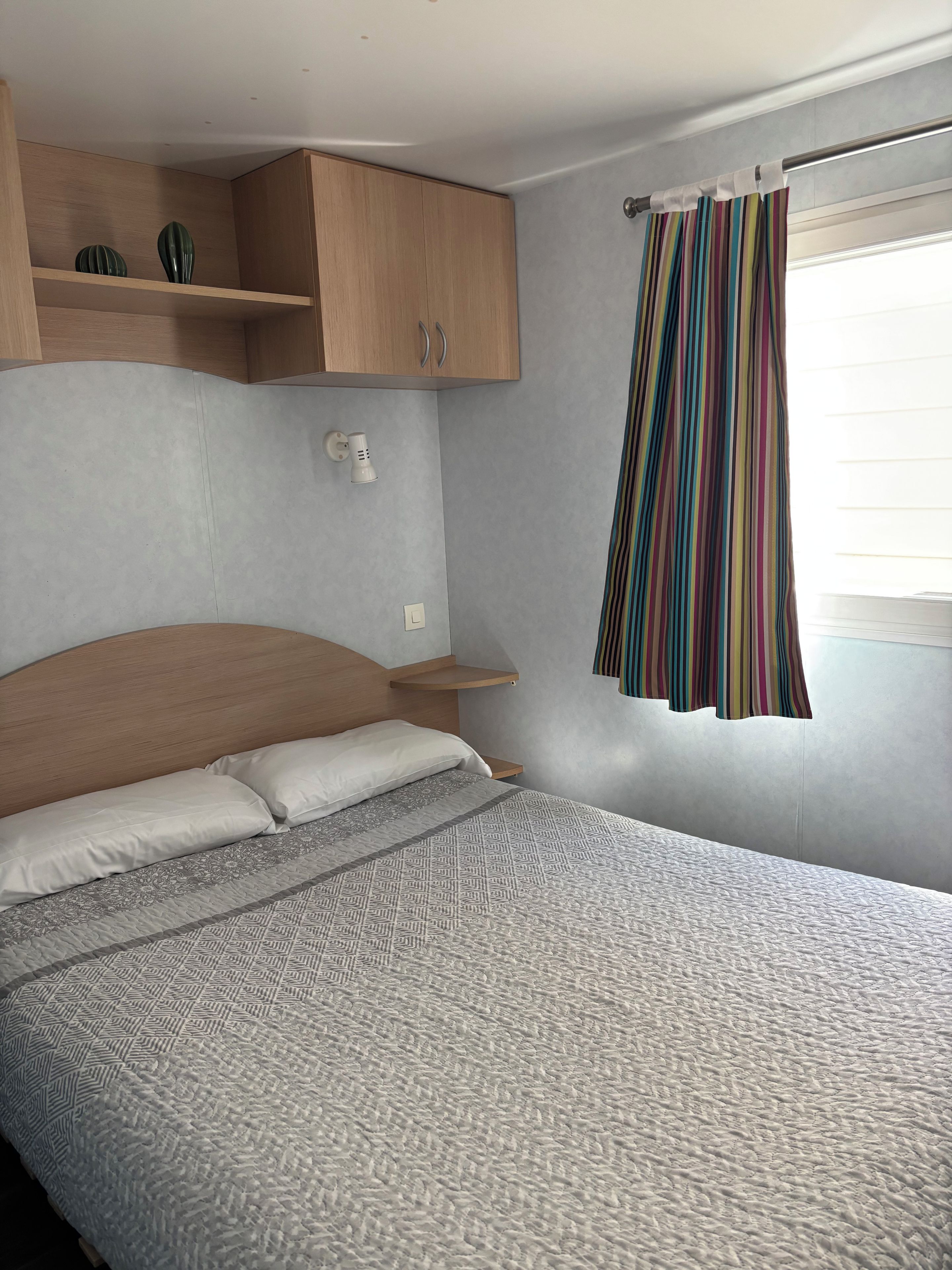 Un dormitorio con una cama, cabecera de madera, mesita de noche, lámpara de pared y cortina a rayas.