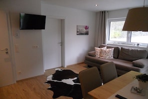 Ein modernes Wohnzimmer mit einem an der Wand montierten Flachbildfernseher, einer Couch mit gemusterten Kissen, einem Esstisch mit Stühlen und einem großen Fenster, das für natürliches Licht sorgt.