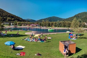 Una zona junto al lago con gente descansando sobre el césped, una pequeña estructura de madera y un bote inflable verde.