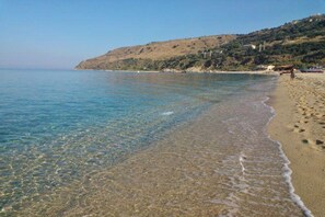 Una playa con aguas claras y poco profundas, una orilla arenosa y un paisaje montañoso al fondo.