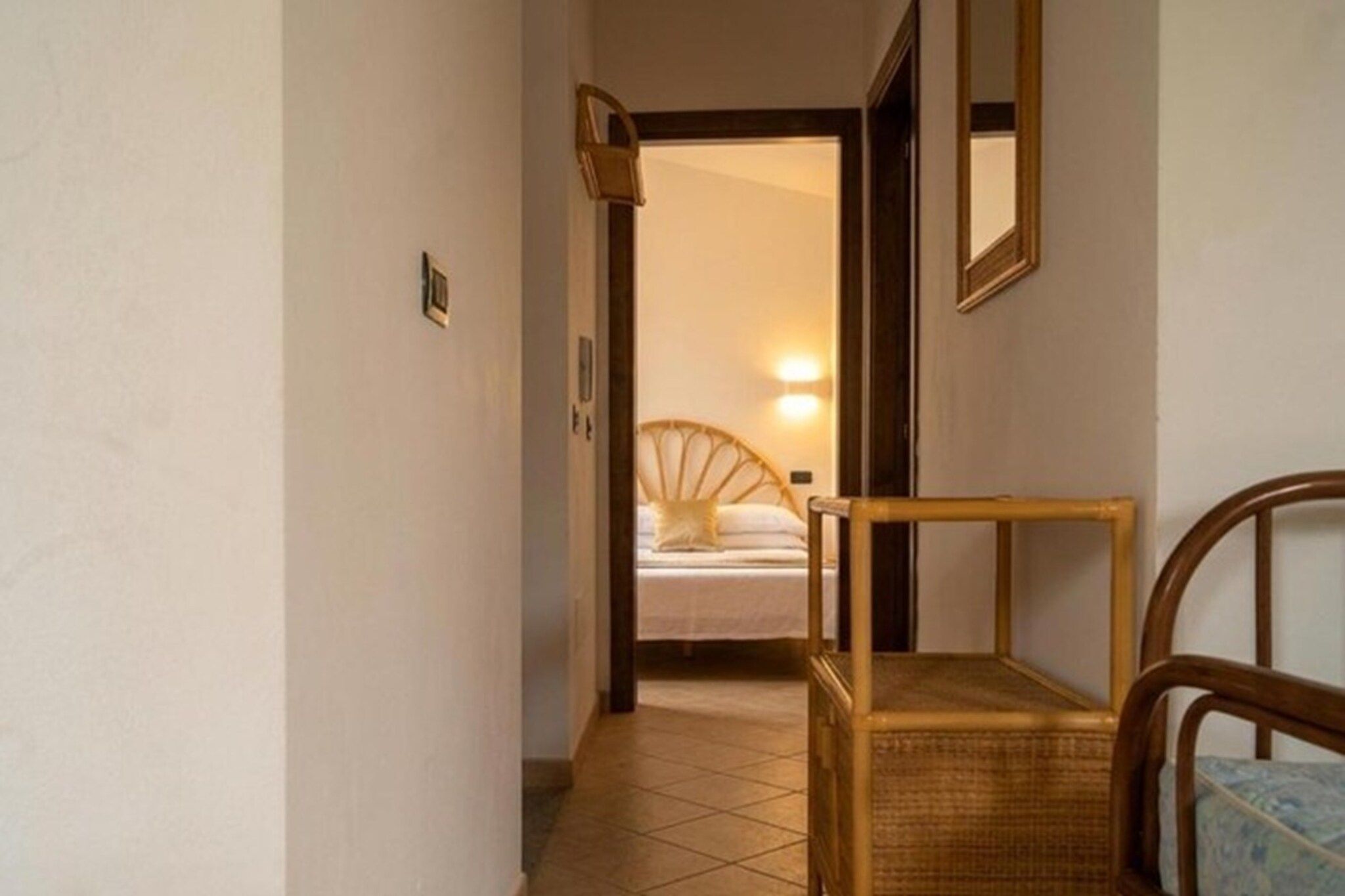 Un pasillo que conduce a un dormitorio con cama y mesita de noche. Hay una silla en primer plano y un espejo en la pared.