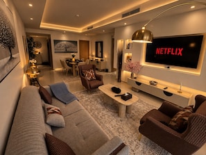 A modern living room with a large flat-screen TV, a sofa, a coffee table, and a dining area in the background. - Burj viewsFrom bed paramount Luxe 3BR+ Pool infinity (Dubai)