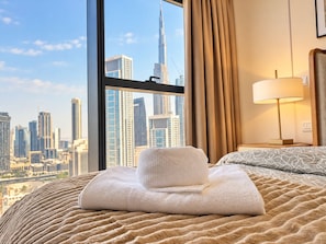 A hotel room with a large window, a bed with a textured blanket, a folded towel, and a bedside lamp. - Burj viewsFrom bed paramount Luxe 3BR+ Pool infinity (Dubai)