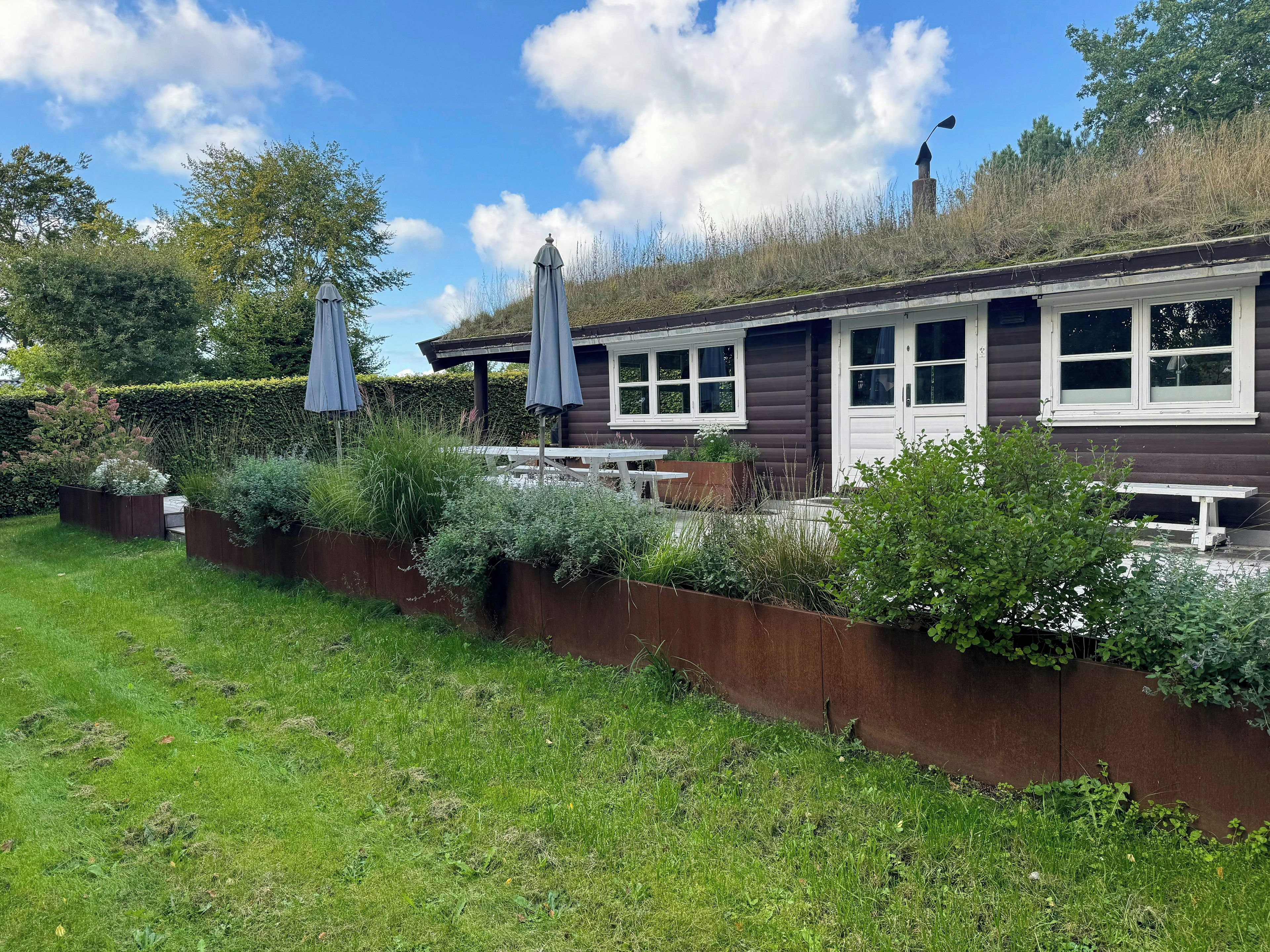 A wooden cabin with a patio area, surrounded by a garden with various plants and a bench.