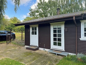Una cabaña de madera con puerta y ventanas blancas, una chimenea y un acceso pavimentado.