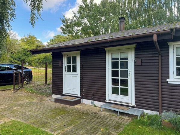 A wooden cabin with a white door and windows, a chimney, and a paved entrance area.