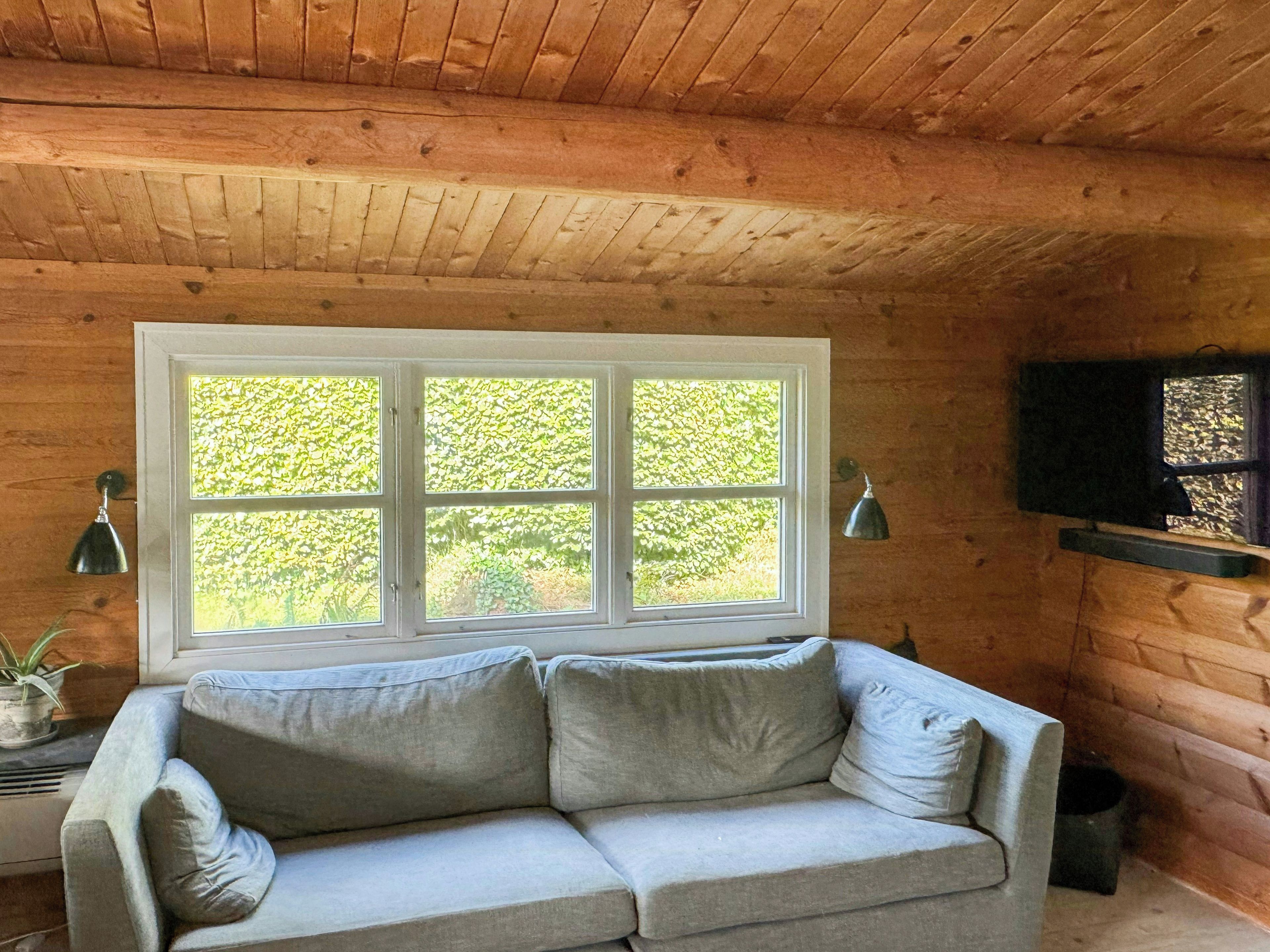 Interior de una cabaña de madera con sofá, ventana y televisor montado.