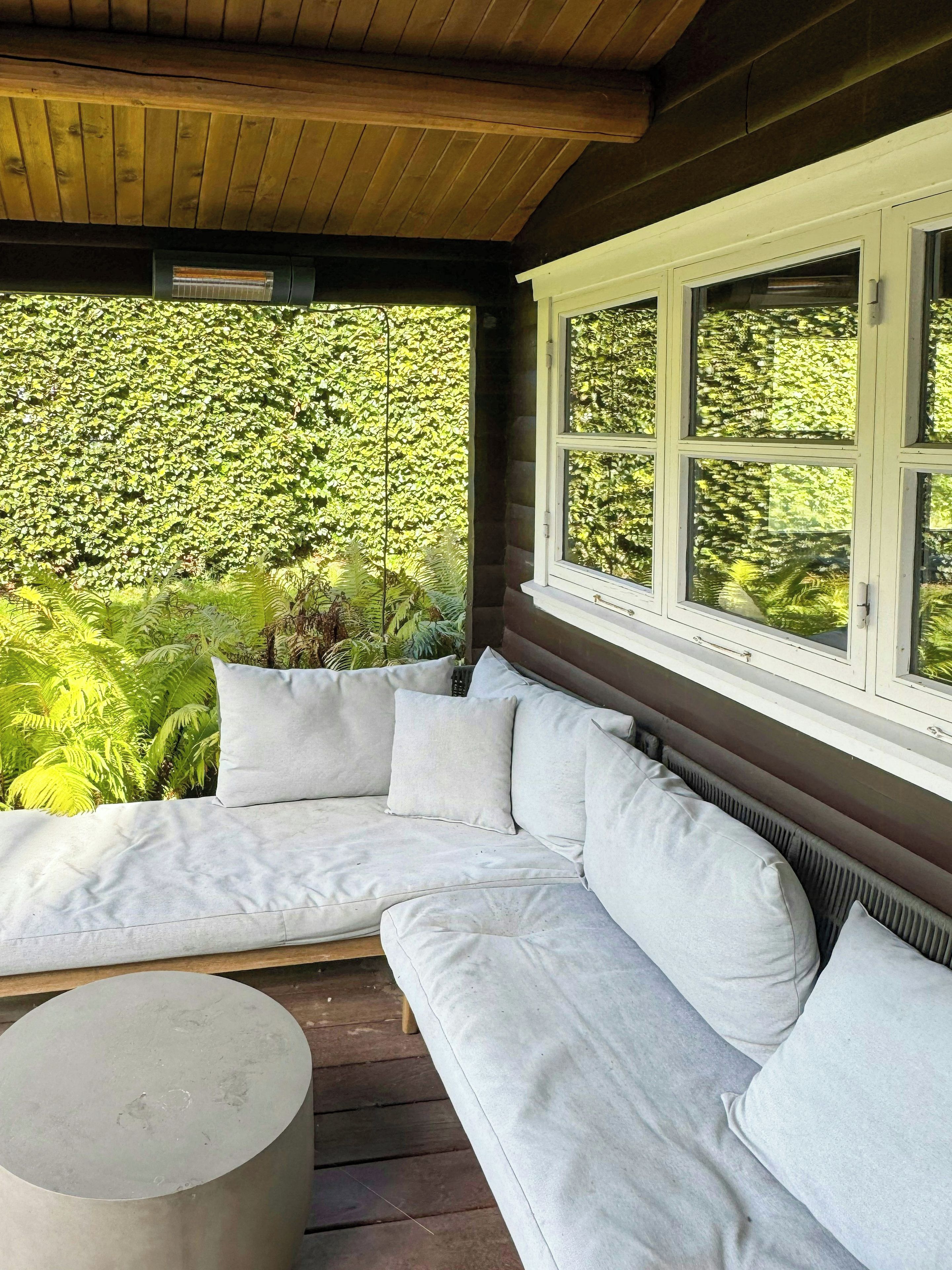 A covered outdoor seating area with a white sofa and a round table, surrounded by lush greenery.