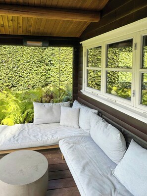 A covered outdoor seating area with a white sofa and a round table, surrounded by lush greenery.