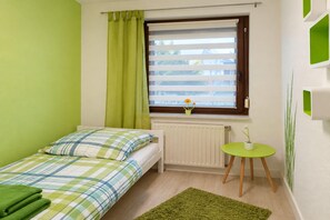 Une petite chambre avec un lit simple, une fenêtre, un radiateur, un tapis vert et une petite table sur laquelle est posée une plante.