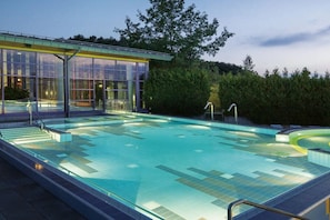 Ein Schwimmbecken mit Glasüberdachung, umgeben von Grünflächen und einem Gebäude mit großen Fenstern.