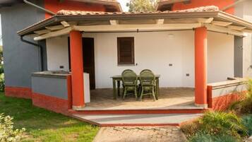 Un espace extérieur abrité, avec deux chaises et une table, une petite fenêtre et une porte donnant accès à un bâtiment.