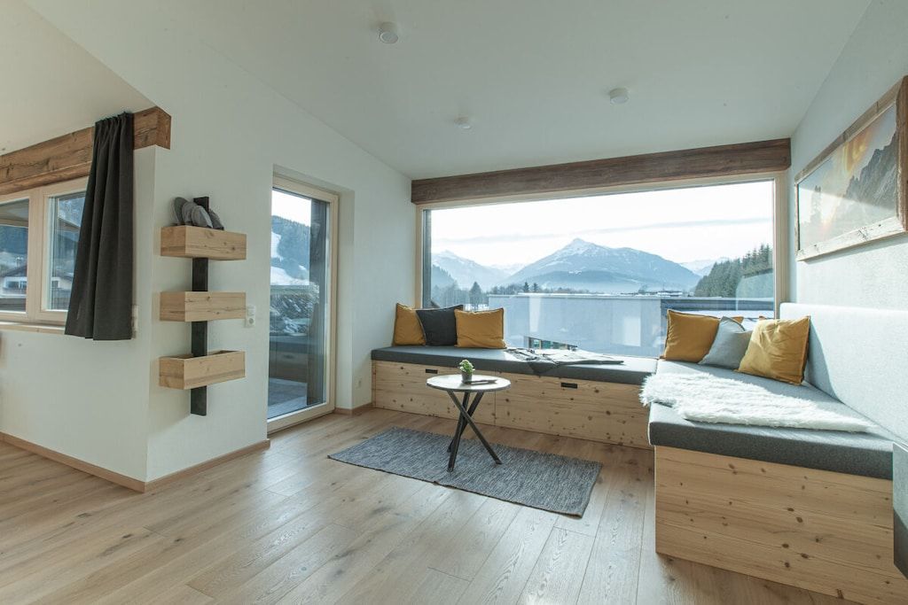 Ein modernes Zimmer mit Holzboden, einer eingebauten Bank mit Kissen, einem kleinen Tisch und einem großen Fenster mit Blick auf die Berge.