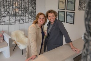 Zwei Personen stehen in einem Raum mit einer weißen Wand, einem weißen Stuhl und gerahmten Karten.