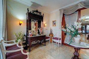 Una habitación con piso de mármol, un mueble de madera con objetos decorativos, un espejo, una silla blanca y una mesa redonda con un jarrón de flores.