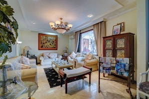 A well-lit living room with a chandelier, a large painting, and a cabinet with magazines. - Cassa Dharmawangsa By ARBA (Jakarta)