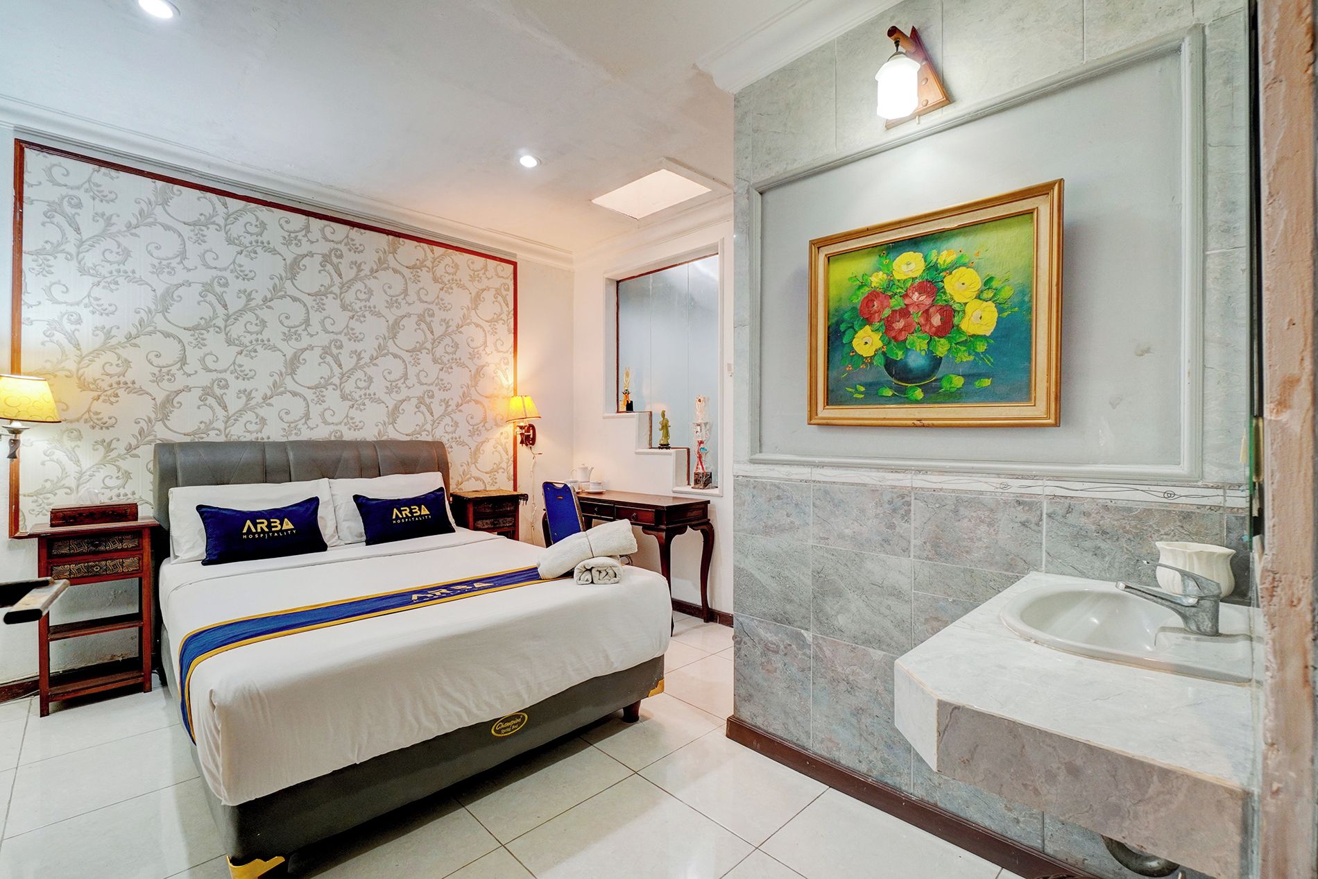 Habitación de hotel con cama, mesitas de noche, lámpara, un cuadro y baño con lavamanos y espejo.