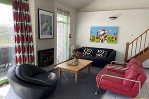 Un salon avec un fauteuil en cuir noir, un canapé rouge, une table basse en bois ornée de fleurs, une cheminée et un tableau représentant des fleurs accroché au mur.