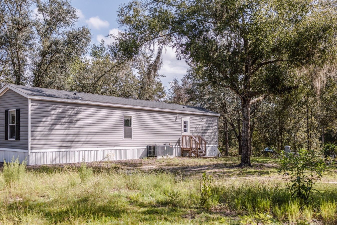 A single-story mobile home with a front porch, surrounded by trees and grass.
