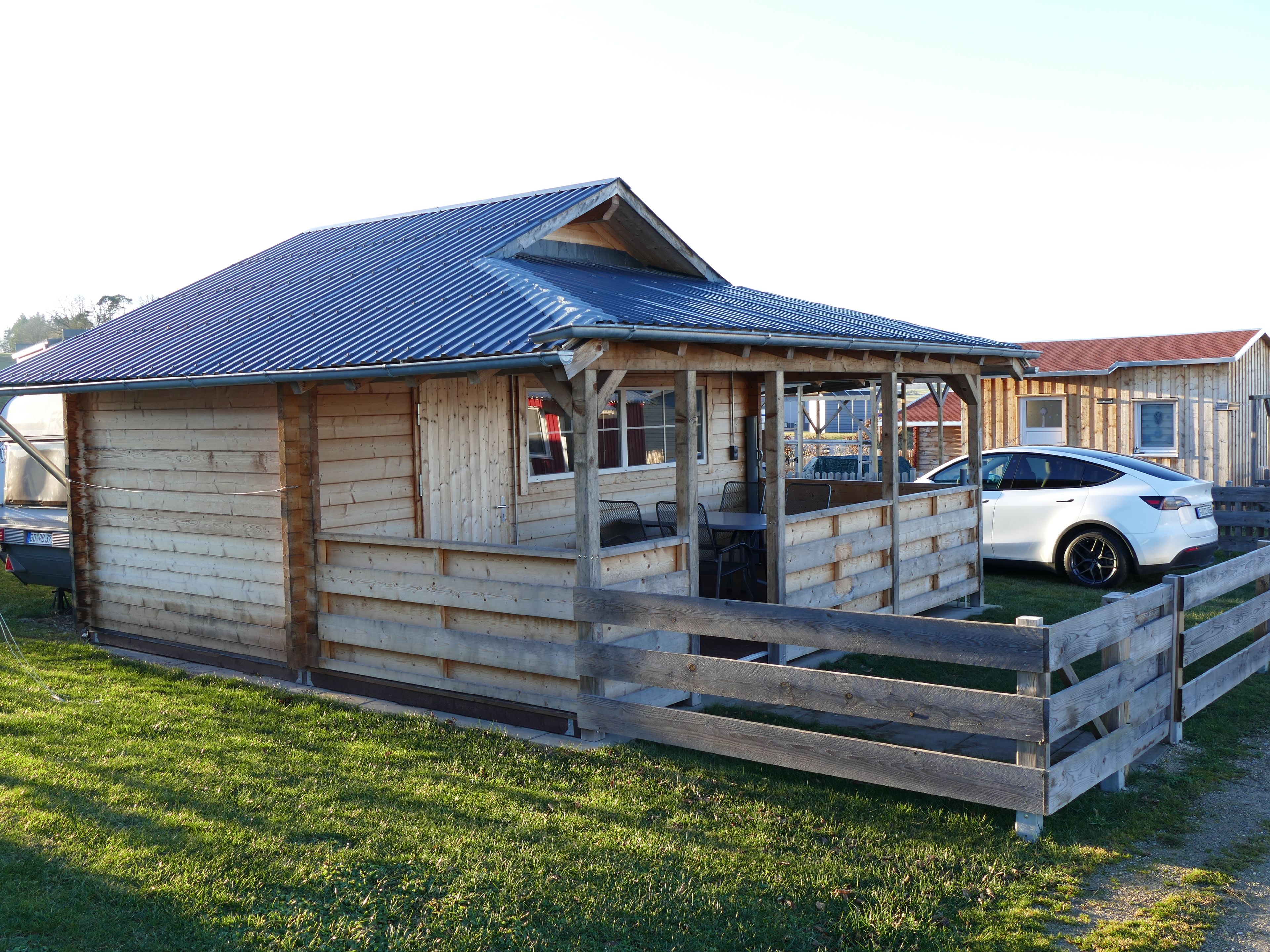 A wooden cabin with a covered porch, a fenced area, and a white car parked nearby.