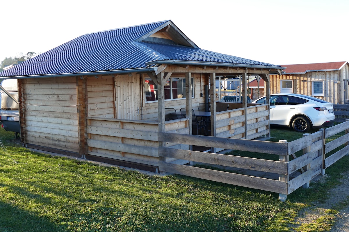 A wooden cabin with a covered porch, a fenced area, and a white car parked nearby.