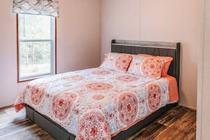 Una cama individual con una colcha estampada y almohadas, una ventana con vistas a árboles, y suelo de madera.