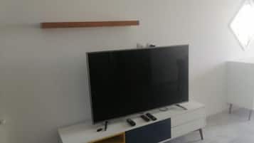 A modern living room with a flat-screen TV on a white cabinet, a wooden shelf above, and a white radiator on the wall.