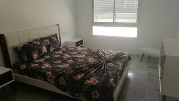 A bedroom with a bed, a bedside table, a nightstand, a window with blinds, and a small white bench.