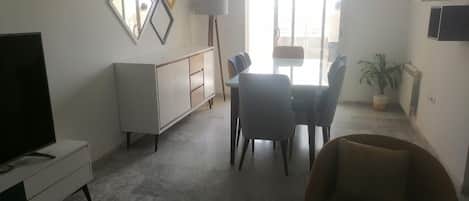 A living room with a television, a dining table with chairs, a sofa, and a potted plant.
