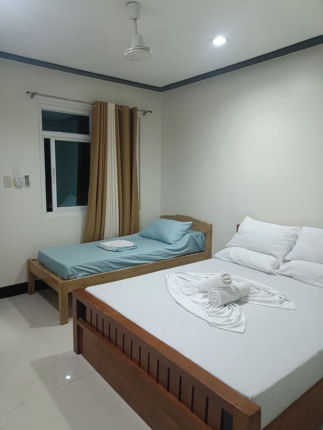 A hotel room with two beds, a window with curtains, a ceiling light, and a thermostat on the wall.