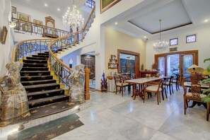 A grand staircase with ornate railings, a chandelier, and a dining area with wooden furniture.