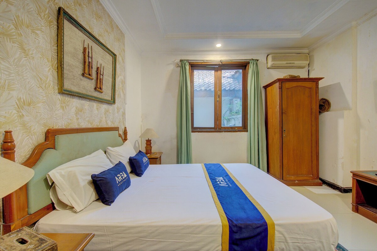 A hotel room with a bed, a wooden wardrobe, a window with curtains, and a wall-mounted air conditioner.