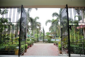 A gated entrance to a property with a paved pathway leading through a garden with various plants and a building in the background. - Shophori Wellness Retreat (Bolpur)