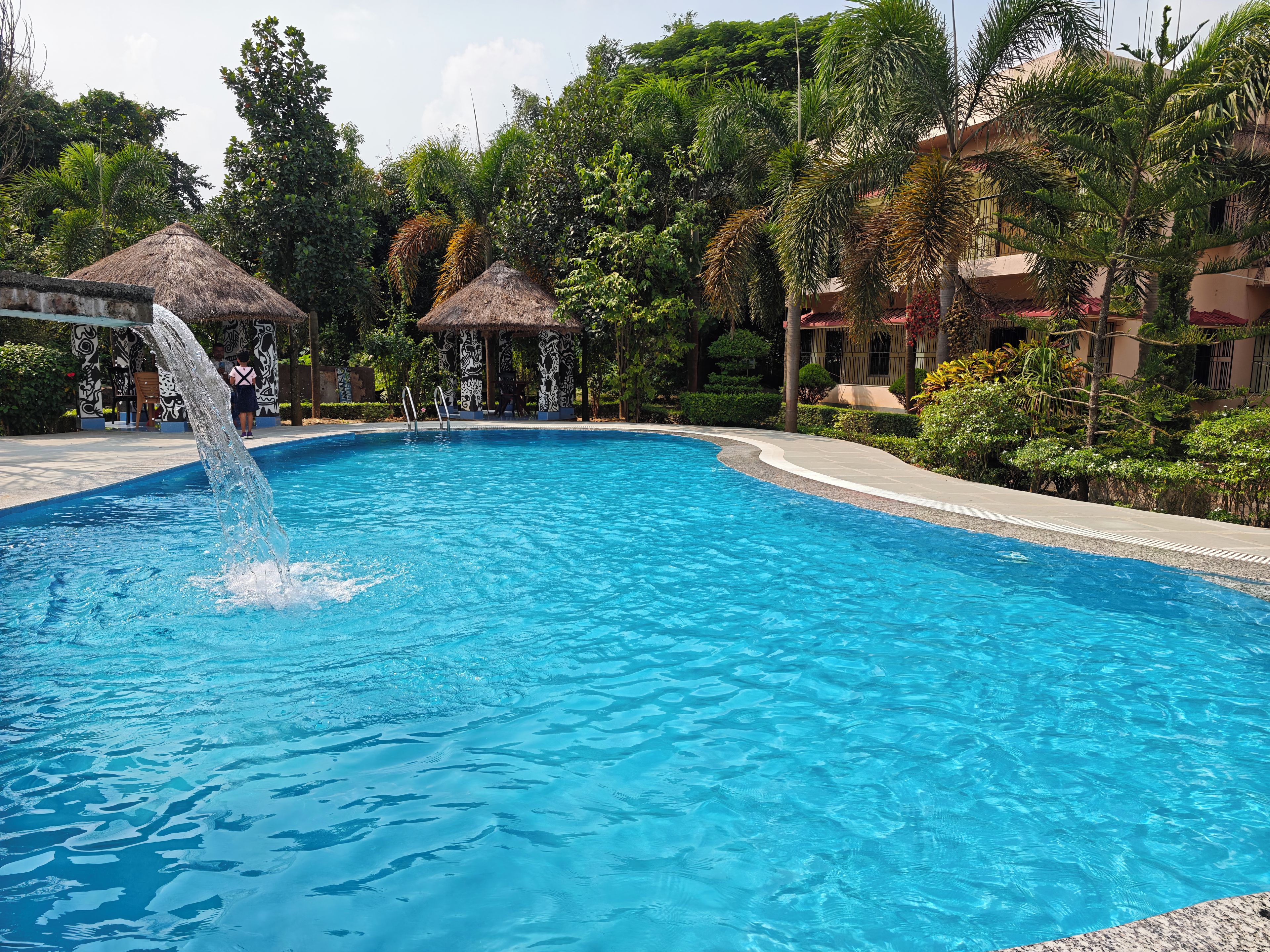 A swimming pool with a water feature, surrounded by lush greenery and buildings.