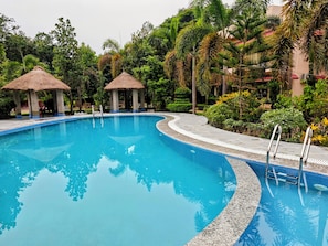 A swimming pool with a curved edge, surrounded by tropical vegetation and two thatched-roof structures.