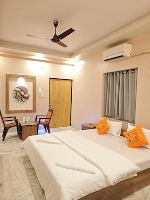 A hotel room with a bed, a desk, chairs, a ceiling fan, an air conditioning unit, a door, and a window with curtains.