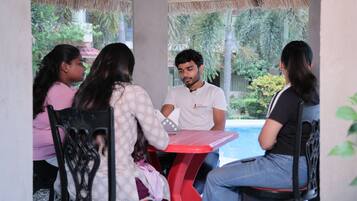 A group of people seated around a red table in an outdoor setting with a pool and greenery in the background.
