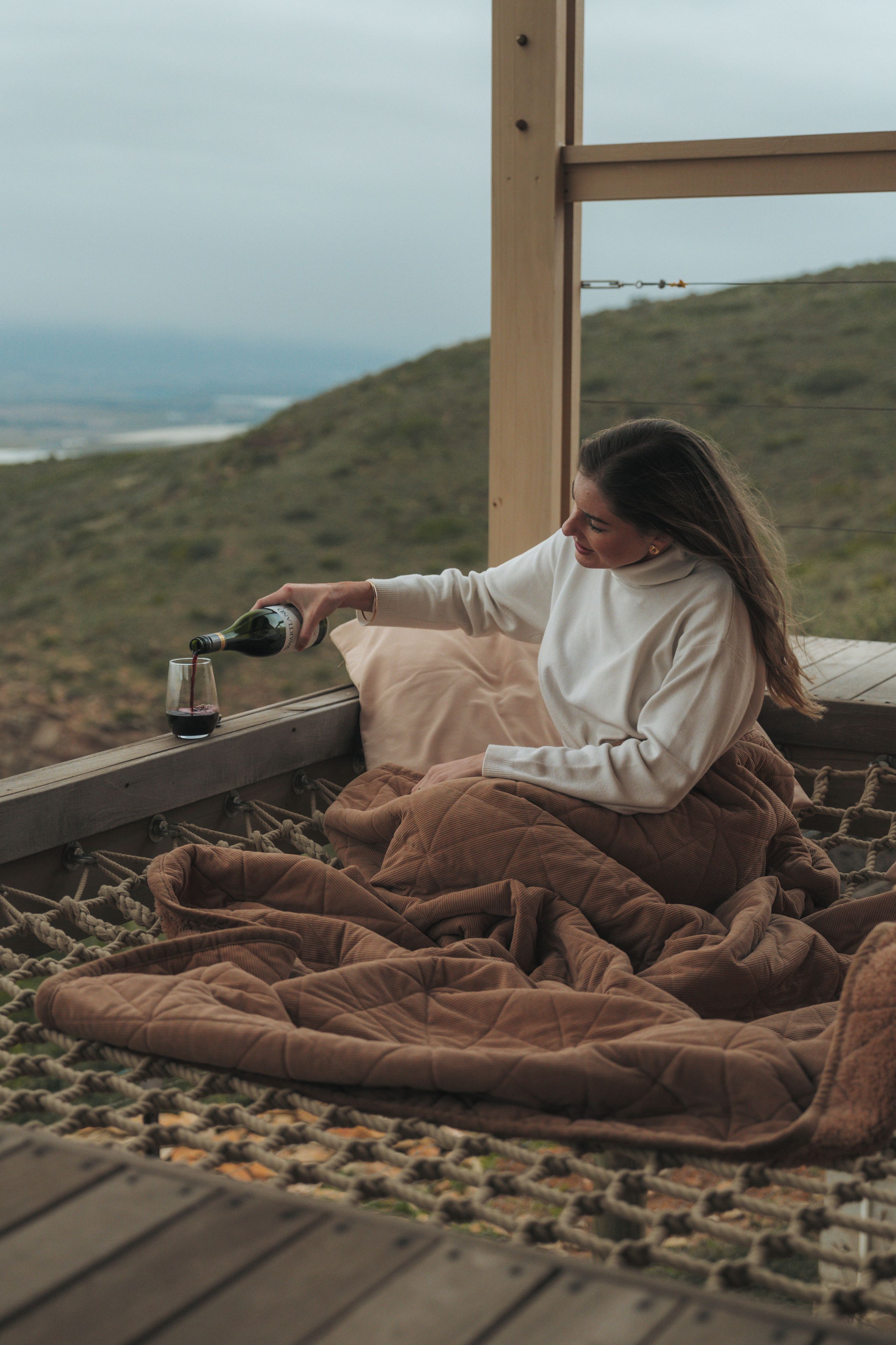 Una persona sirve vino en una copa sobre una terraza de madera con vistas a un paisaje montañoso.