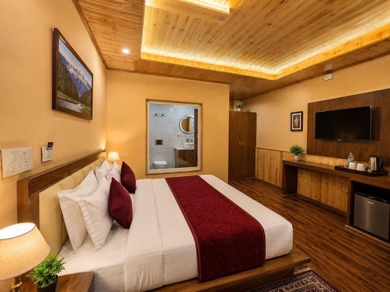 A hotel room with a bed, a television, a desk, and a bathroom visible through a glass door.