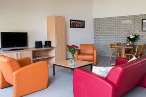 Ein modernes Wohnzimmer mit Flachbildfernseher, orangefarbenen und roten Sofas, einem Holzkabinett und einem Essbereich mit Backsteinwand.
