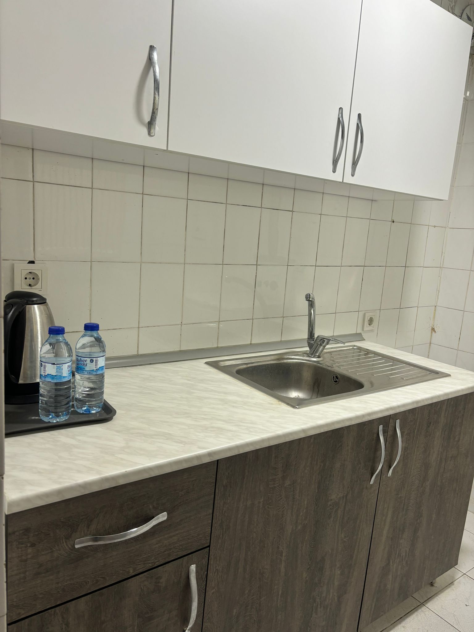 Una cocina con un lavamanos, dos botellas de agua, una tetera y armarios.