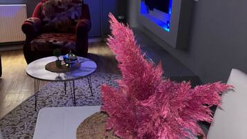 Una sala de estar con un sofá blanco, una mesa de centro, un florero decorativo con follaje rosa, un sillón rojo y un televisor montado en la pared con retroiluminación azul.