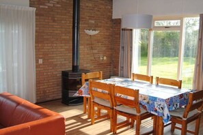 A dining area with a table set for four, wooden chairs, a brick wall, a chimney, and a view of a green landscape through large windows. - De Branding 53, Kustlicht, ZOUTELANDE (ZOUTELANDE)