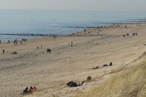 A beach with people walking and sitting on the sand, a wooden pier in the background, and a grassy area on the right. - De Branding 53, Kustlicht, ZOUTELANDE (ZOUTELANDE)